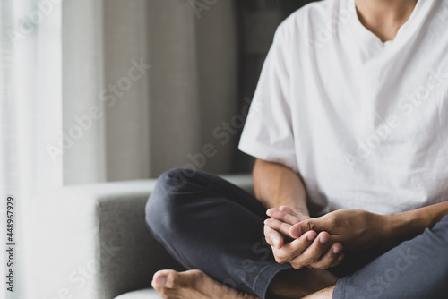 Young man in a white t-shirt and grey pants sat on the sofa practicing meditation  sitting in a lotus pose alone in the morning at home. During the quarantine due to the spread of the coronavirus.