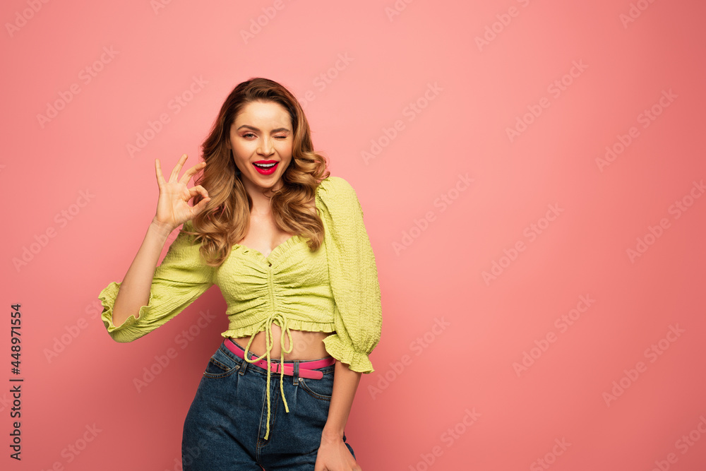 cheerful woman in green blouse showing ok sign while winking eye isolated on pink cheerful woman in green blouse showing ok sign while winking eye isolated on pink
