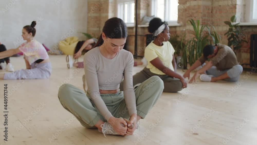 Selective focus shot of young woman in sportswear doing seated butterfly stretch while preparing with team of multiethnic dancers for dance class in studio Selective focus shot of young woman in sportswear doing seated butterfly stretch while preparing with team of multiethnic dancers for dance class in studio