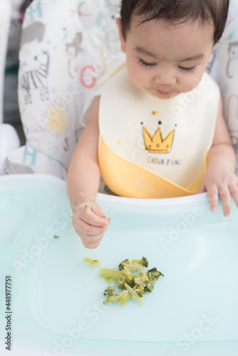 Top view of cute Asian baby girl wearing  baby apron sitting on dining table eat broccoli by her self, Baby Led Weaning concept