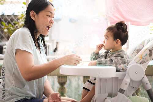 Side view of Asian mother try to practice her cute baby girl eat food by her self, she wearing baby apron sitting on dining table , Baby-Led Weaning concept
