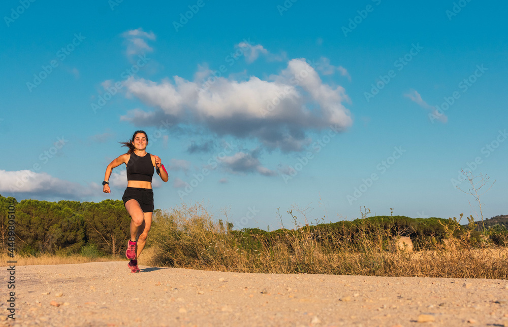 Front view of a caucasian young woman running and smiling in a mountain path during the golden hour. Front view of a caucasian young woman running and smiling in a mountain path during the golden hour.