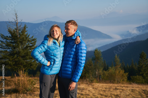 Two happy hikers man and woman standing and embracing on the background of trees, mountain hills, covered by forests and silhouettes mountain peaks far away. Active hiking in the mountains. © anatoliy_gleb