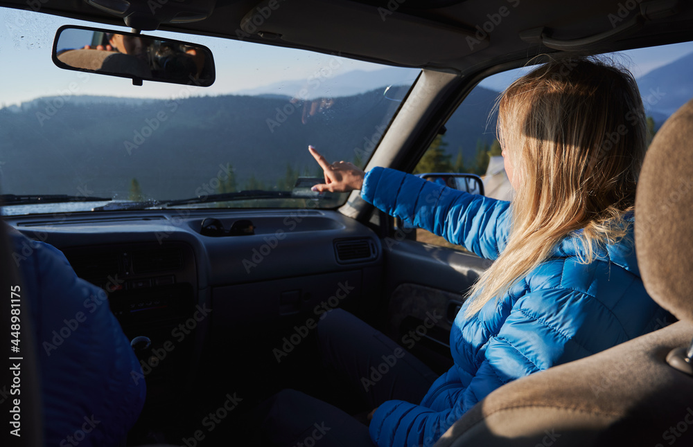 Young woman sitting on passenger seat in car and pointing finger at hills while travelling with husband. Female traveler sitting in automobile next to driver and gesturing towards beautiful mountains.