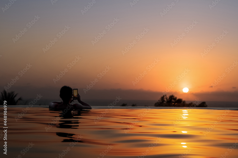 A young woman in the outdoor pool of the hotel photographs a beautiful sea sunset by smartphone. Silhouette of a girl on the background of sunset. Rest at the sea.