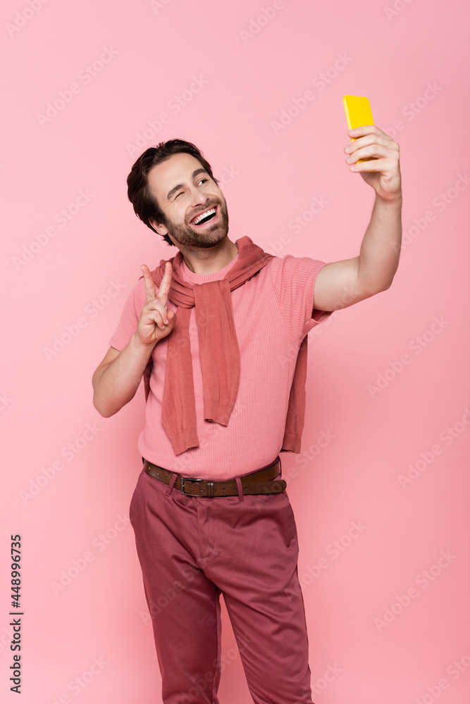 Smiling man showing victory gesture while taking selfie on smartphone isolated on pink Smiling man showing victory gesture while taking selfie on smartphone isolated on pink