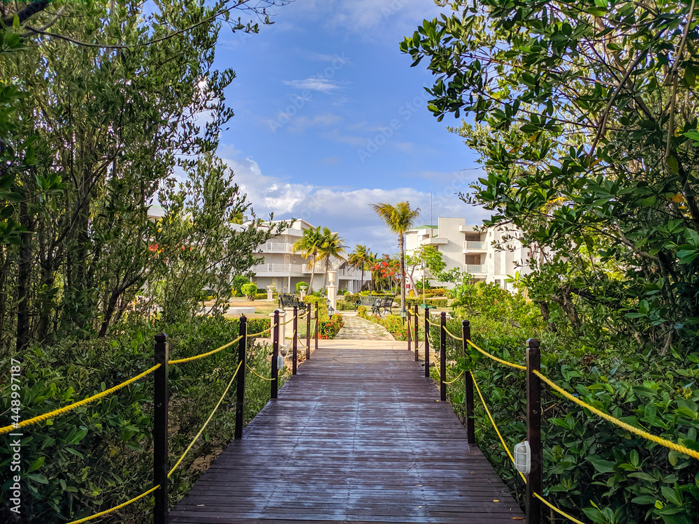 View from the wooden bridge surrounded by the jungle to the hotel Tryp Cayo Coco grounds. From the bridge you can see the hotel's white buildings and a seating area with benches for guests.