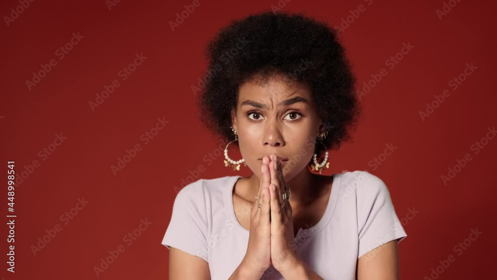 A worried african american woman holding hands in a pray gesture while standing isolated over red wall in the studio A worried african american woman holding hands in a pray gesture while standing isolated over red wall in the studio