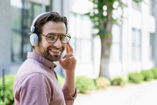 Smiling businessman in eyeglasses and headphones looking at camera outdoors