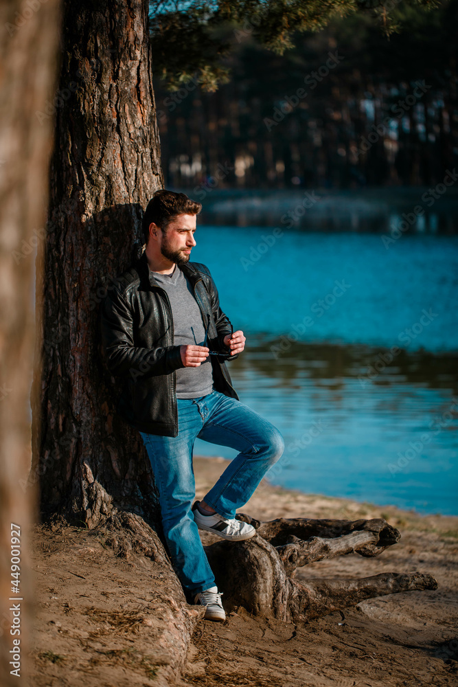 Young man in a leather jacket stands pensive and serious under a tree in autumn Young man in a leather jacket stands pensive and serious under a tree in autumn