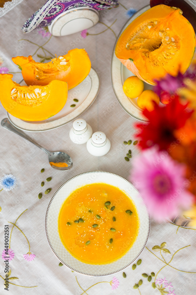 Plate with colorful pumpkin soup, pumpkin cut into pieces on porcelain plates, pepper shaker, salt shaker, spoon, red gerbera flower and green pumpkin seeds on white tablecloth Plate with colorful pumpkin soup, pumpkin cut into pieces on porcelain plates, pepper shaker, salt shaker, spoon, red gerbera flower and green pumpkin seeds on white tablecloth