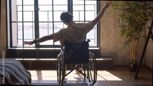 Rear view backlighted of man sitting in wheelchair in apartment, wearing over-ear headphones, listening to music and moving hands photo