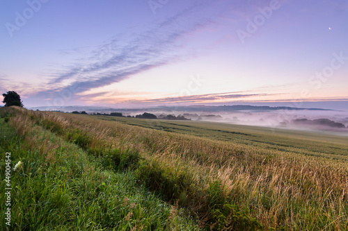 Morning fog under the agricultural field. Summertime rural landscape with fields, hills and beautiful sky. Sunrise in the village