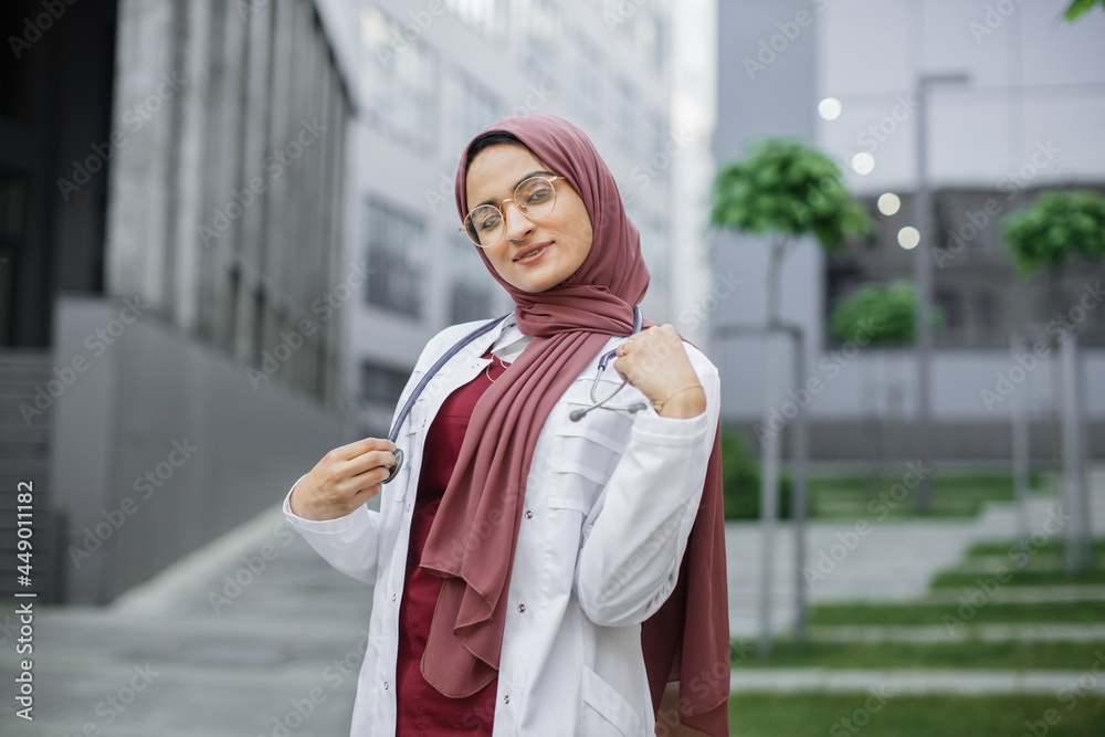 Beautiful Malay lady doctor wearing hijab, eyeglasses and stethoscope outside modern clinic. Closeup portrait of friendly, smiling confident muslim female doctor in scrubs, standing outdoors