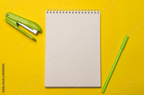 Green stationery stapler, green pen and a clean white sheet of notepad on a yellow background, top view. Back to school.
