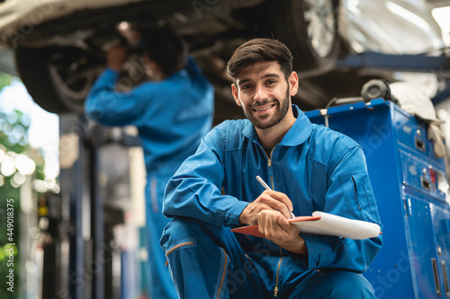 Portrait photo of professional look Caucasian vehicle service technician kneeling in front of car repair shop and holding car inspect check list.