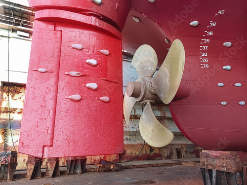 The stern of a large ship with a copper propeller, with a steering gear on a slipway in a dry dock. Dry-cargo ship in dry dock for repairs and maintenance at a shipyard