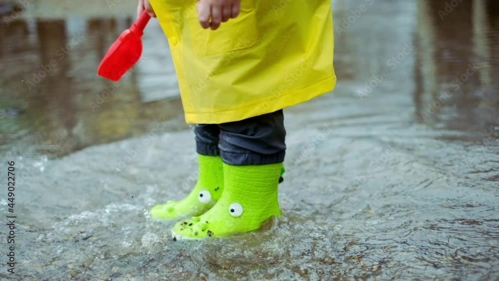 child in green rubber boots and yellow raincoat is jumping in puddle in ...