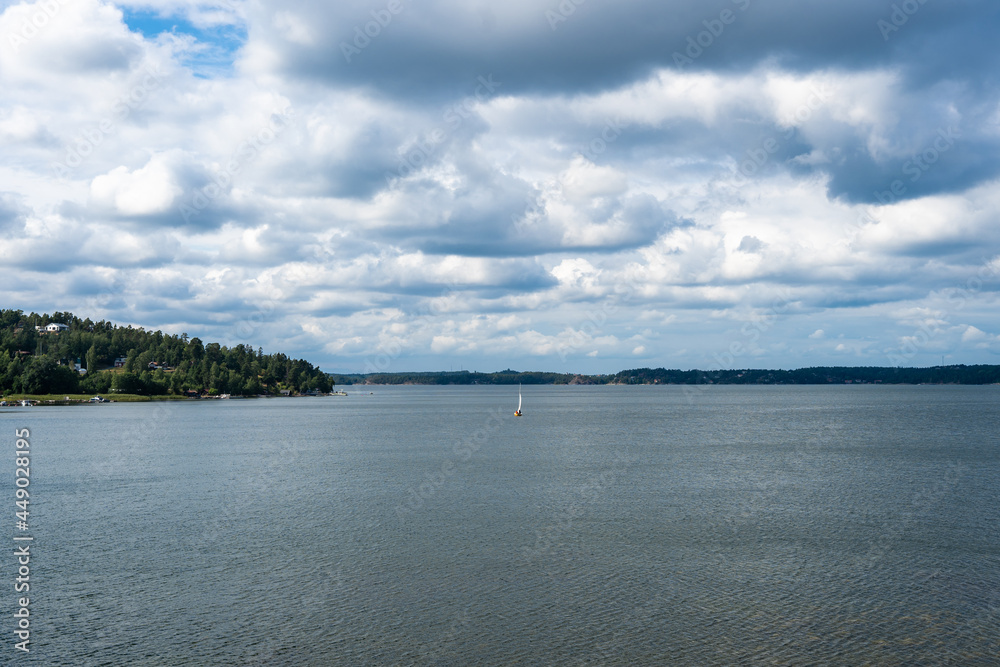 Dramatic sky. Panorama of the Baltic sea bay with white sailing yacht on water surface. Beautiful grey clouds floating on the horizon. Forested islands overgrown with evergreens trees.