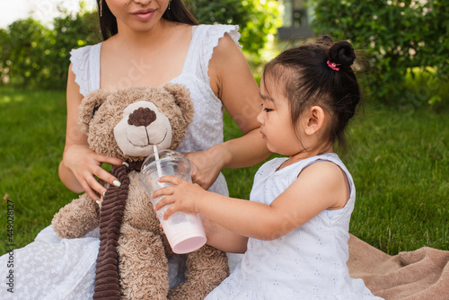 asian toddler girl holding plastic cup with milkshake near teddy bear in hands of mother