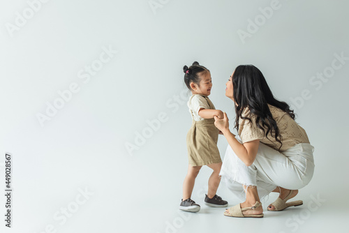 full length of happy asian toddler kid holding hands with stylish mother on grey