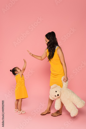 cheerful asian woman holding teddy bear and giving high five to happy toddler daughter on pink