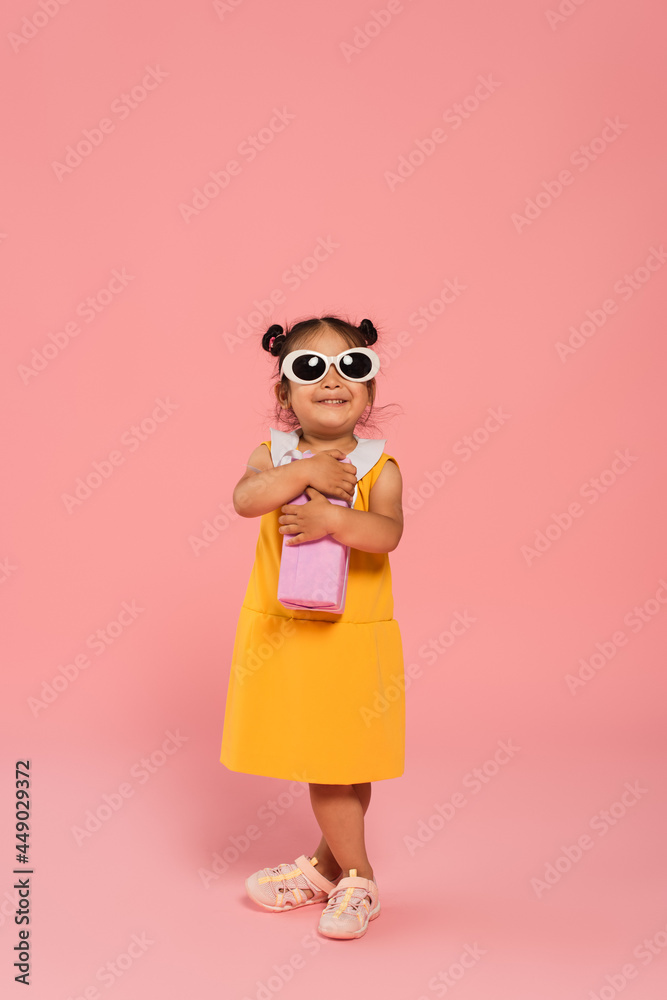 full length of cheerful asian toddler kid in yellow dress and sunglasses holding present on pink full length of cheerful asian toddler kid in yellow dress and sunglasses holding present on pink
