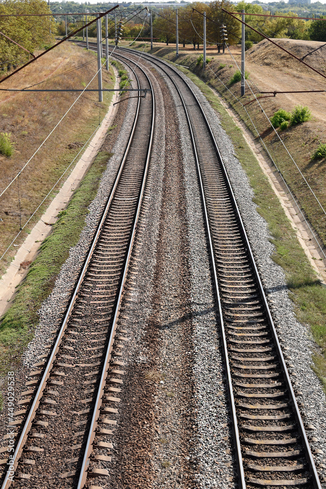 Two railroad tracks lie on a crushed rock railroad bank and lead to the left into the woods. Top view.