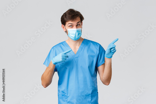 Covid-19, healthcare workers and hospital concept. Doctor inviting look at advertisement, take look. Nurse in medical mask, gloves and scrubs pointing fingers right, showing promo, grey background