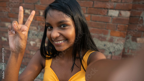 Portrait of a young happy dark-skinned Indian teenage girl posing, taking a selfie by showing peace sign gesture hand, victory gesture towards the camera, smiling genuinely against a brick wall