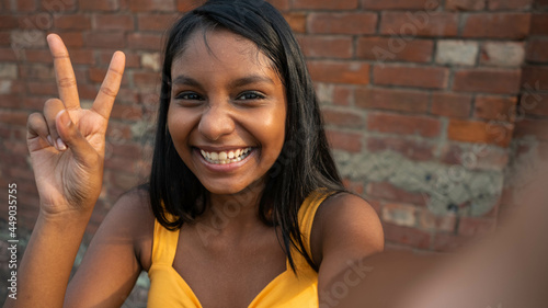 Portrait of a young happy dark-skinned Indian teenage girl posing, taking a selfie by showing peace sign gesture hand, victory gesture towards the camera, laughing genuinely against a brick wall