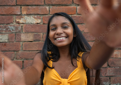 Young brunette girl smiling at the camera with open arms for a hug, reaching out forwards by giving friendly expression, smiling with grin waiting for a hug against a red brick wall