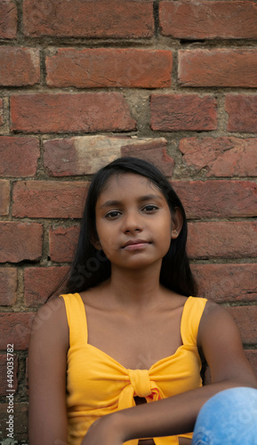 Portrait of a sad, lonely young dark-skinned girl in casual clothes sitting in despair leaning against the red brick wall