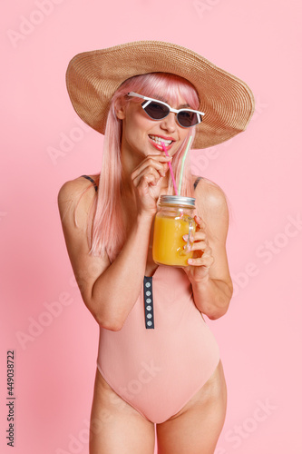 Fashionable woman with pink hair wearing straw hat, swimsuit and sunglasses drinking cocktail, posing isolated over pink studio background. Summer vacation concept