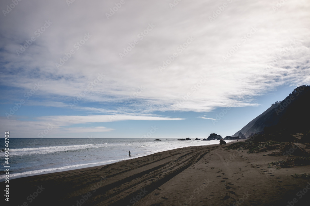 Alone fisher and quad bike in a big and empty beach with hills and blue sky with clouds