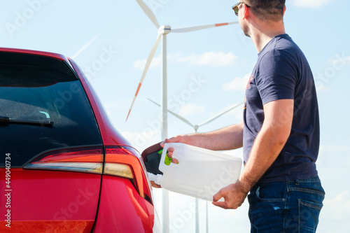 Man filling a diesel engine fluid from canister into the tank of red car on the background of wind turbines. Reduction of air pollution and environmental protection. Green energy concept.