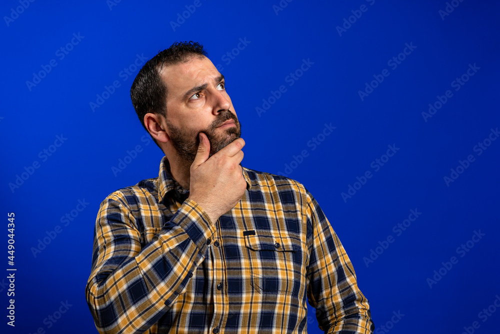 Frowning man thinking expressing doubts and concerns. Hipster male with beard in blue plaid checkered shirt Isolated on blue studio Background. Negative face expression, human emotion, body language. Frowning man thinking expressing doubts and concerns. Hipster male with beard in blue plaid checkered shirt Isolated on blue studio Background. Negative face expression, human emotion, body language.