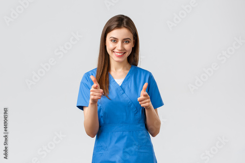 Healthcare workers, prevent virus, insurance and medicine concept. Enthusiastic smiling female nurse, doctor in blue scrubs, pointing herself with pistols, choosing, inviting clients to clinic