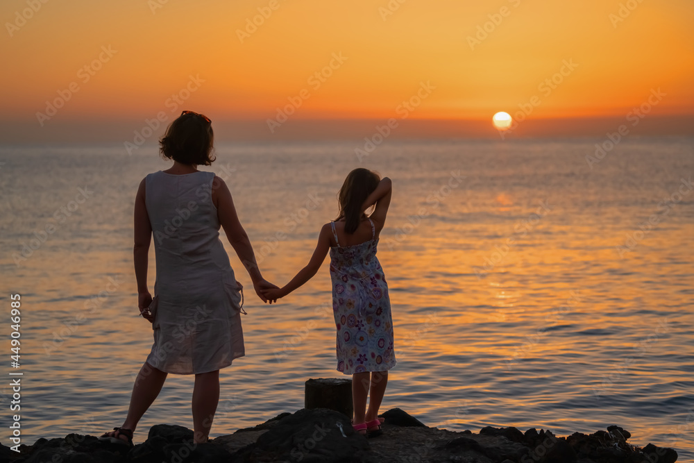 Back view of young mom and daughter are watching the sunrise at sea. Happy family concept.