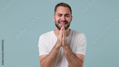 Pleading begging young brunet man 20s wears white t-shirt hold hands folded in prayer making wish keep fingers crossed isolated on pastel light blue background. People emotions lifestyle concept