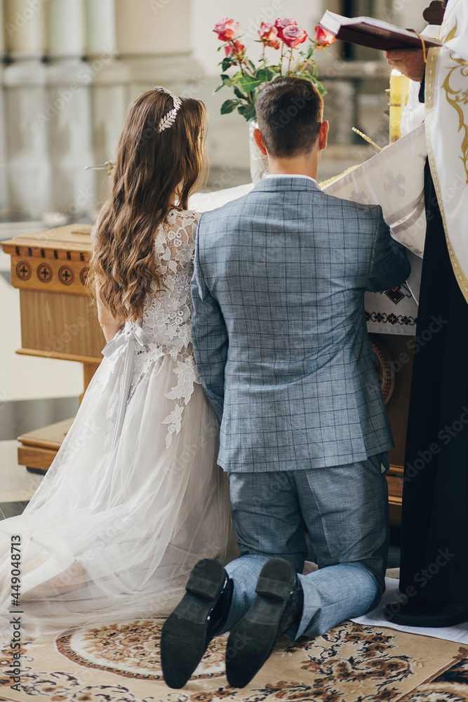 Stylish emotional bride and groom making oaths and holding hands on holy bible during matrimony in church. Wedding ceremony in cathedral. Classic wedding couple giving vows. Back view