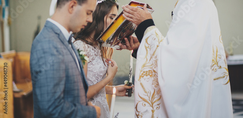 Stylish bride and groom kissing holy bible from priest hands during matrimony in church. Wedding ceremony in cathedral. Classic spiritual wedding couple