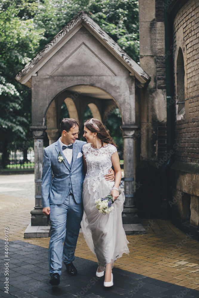 Stylish happy bride and groom walking on background of church in sunny street. Provence wedding. Beautiful emotional wedding couple smiling and walking in european city.