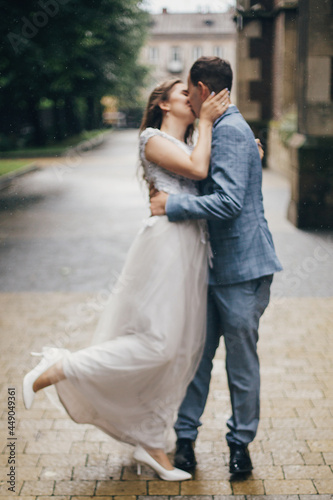 Blurred image of stylish sensual bride and groom kissing on background of old church in rain. Provence wedding. Beautiful emotional wedding couple embracing. Romantic moment