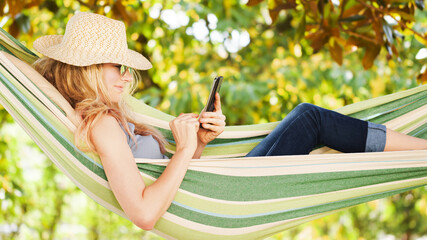 Smiling blonde woman with sunglasses using smartphone, lying relaxing on the hammock in the garden, free time and summer holiday concept to surf the internet or chat with friends using social media
