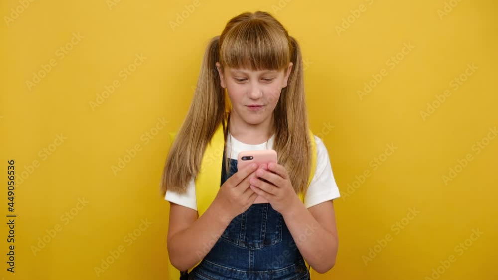 Smiling schoolgirl with backpack holding smartphone enjoying using mobile apps, playing video games, child learning in cellphone, having fun with mobile technology concept, posing on yellow background