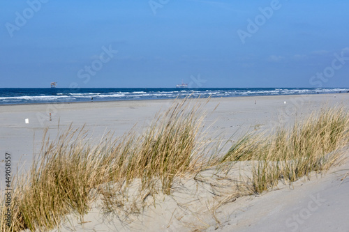 Fototapeta Naklejka Na Ścianę i Meble -  Ameland,Netherlands April 20,2021-Beach with offshore platform, sand, beach grass and surf. People walking on the beach. NAM, Oil rig. Natural gas extraction in the Wadden-North Sea Region.