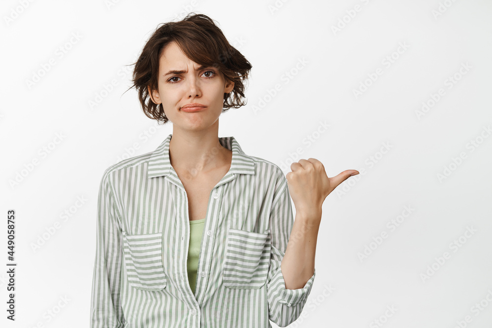 Skeptical young woman frowning, making disappointed smirk while pointing right, showing something upsetting, standing against white background Skeptical young woman frowning, making disappointed smirk while pointing right, showing something upsetting, standing against white background