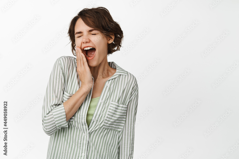 Tired cute brunette girl yawning, feeling fatigue, sleepy face, covering open mouth with hand, standing against white background Tired cute brunette girl yawning, feeling fatigue, sleepy face, covering open mouth with hand, standing against white background