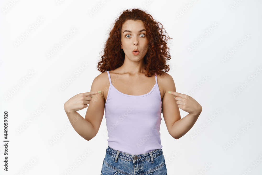 Surprised redhead woman say wow, pointing finger at herself with amazed face, achieve goal, winning smth, standing in tank top against white background Surprised redhead woman say wow, pointing finger at herself with amazed face, achieve goal, winning smth, standing in tank top against white background